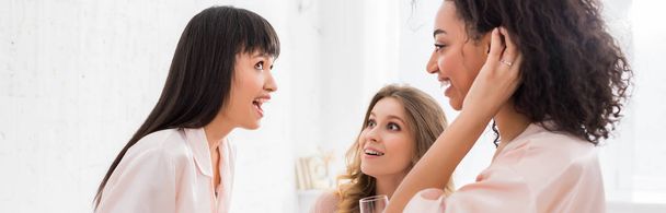 tiro panorâmico de namoradas multiétnicas excitadas com copos de champanhe falando na festa de pijama
 - Foto, Imagem