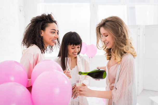 alegre multicultural niñas verter champán de botella en vasos en despedida de soltera con globos de color rosa
 - Foto, Imagen