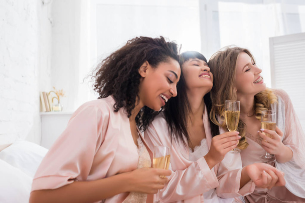 usmívající se multietnické přítelkyně baví a drží sklenice šampaňského na pyžamové párty - Fotografie, Obrázek