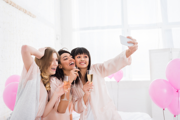 attraenti ragazze multiculturali con bicchieri di champagne scattare selfie su smartphone durante il pigiama party
 - Foto, immagini