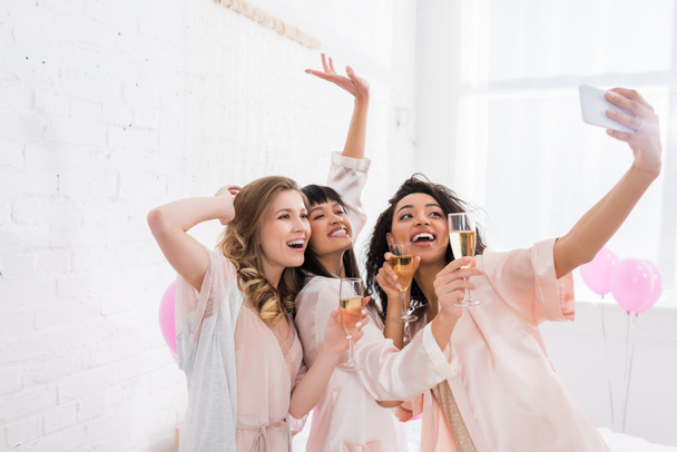 excited multicultural girls with glasses of champagne taking selfie on smartphone during pajama party - Photo, Image