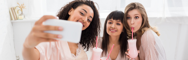 πανοραμική λήψη χαρούμενων πολυπολιτισμικών φιλενάδων σε μπουρνούζια πίνοντας milkshakes ενώ βγάζατε selfie στο smartphone κατά τη διάρκεια bachelorette πάρτι - Φωτογραφία, εικόνα