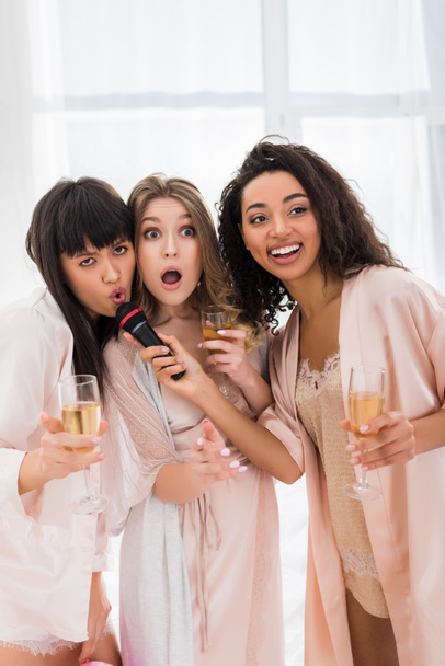красивые возбужденные мультикультурные девушки, держащие шампанское и поющие с микрофоном на девичнике
 - Фото, изображение