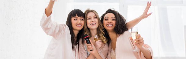 panoramatický záběr atraktivních veselých multikulturních dívek držících šampaňské a zpívajících s mikrofonem na rozlučce se svobodou - Fotografie, Obrázek