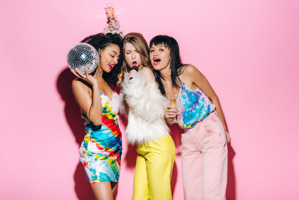 emotional multiethnic girls holding champagne glasses and disco ball while singing with microphone on pink  - Foto, Bild
