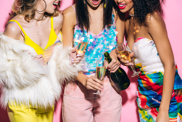 cropped view of emotional multicultural girlfriends pouring champagne from bottle into glasses on pink - Fotó, kép