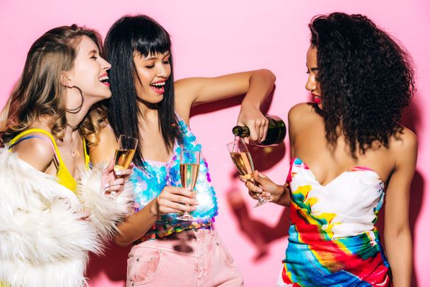 emotional multicultural girlfriends pouring champagne from bottle into glasses on pink - Foto, immagini