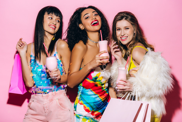 excited fashionable multiethnic girls posing with milkshakes and shopping bags on pink - Foto, Imagem