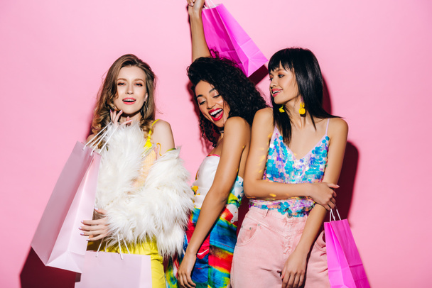 excited fashionable multiethnic girls posing with shopping bags on pink - Photo, Image