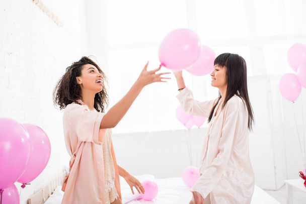 chicas emocionales multiculturales en albornoces sosteniendo globos rosados en despedida de soltera
 - Foto, Imagen