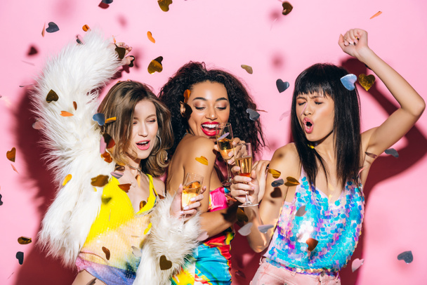 ενθουσιασμένοι μοντέρνα πολυπολιτισμικά κορίτσια χορεύουν με ποτήρια σαμπάνιας σε ροζ με κομφετί - Φωτογραφία, εικόνα