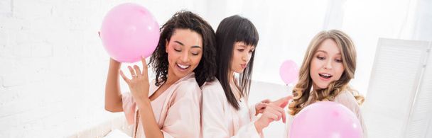 panoramic shot of multicultural girls having fun with pink balloons in bedroom - Photo, Image