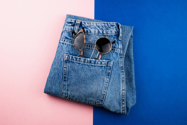Новые джинсы в унисекс и две пары солнцезащитных очков лежат на красочном розовом и модном голубом фоне. Ежедневный минимальный стиль одежды макет сверху вид. Шаблон для веб, рекламы, социальных сетей
. - Фото, изображение