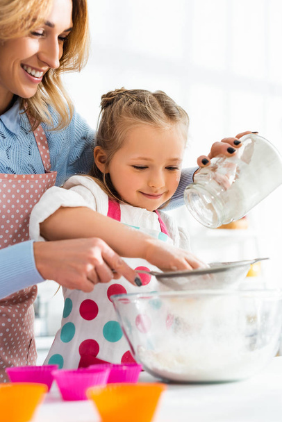 Selective focus of beautiful mother and cute daughter sifting flour through a sieve into bowl  - Fotó, kép