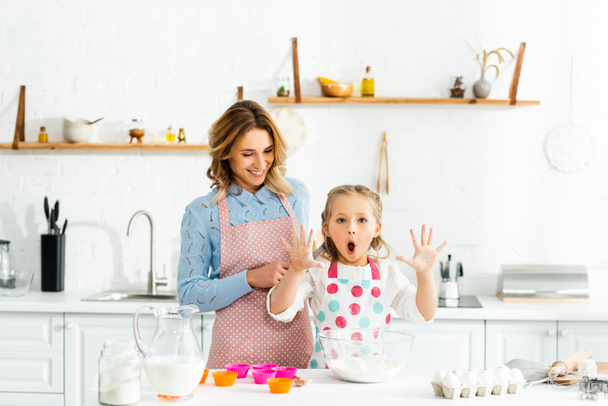 Cute daughter with open mouth holding up hands standing with mom at table  - Photo, Image