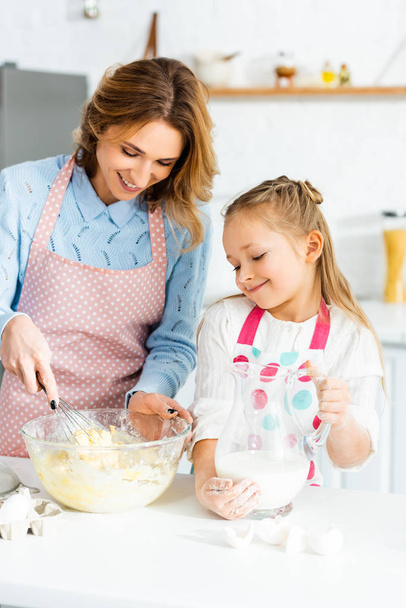 χαμογελαστή μητέρα μαγείρεμα με σύρμα μπαλόνι και η κόρη κρατώντας κανάτα με γάλα  - Φωτογραφία, εικόνα