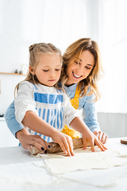 Улыбающаяся мать и милая дочь катят тесто на стол
  - Фото, изображение