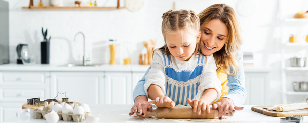 panoramic shot of smiling mother and cute daughter rolling dough on table  - Foto, immagini