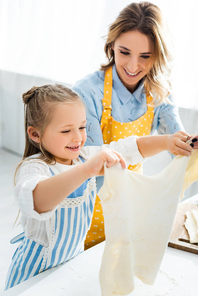 smiling mother looking at cute daughter with dough in kitchen  - Фото, изображение