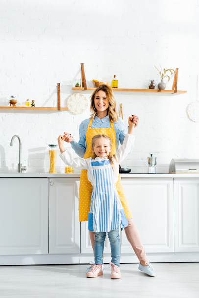 smiling mother holding hands with daughter and looking at camera in kitchen  - Φωτογραφία, εικόνα