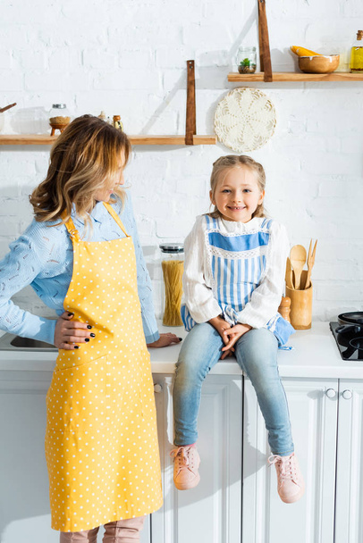 Mutter in Schürze schaut lächelnde und süße Tochter in der Küche an  - Foto, Bild
