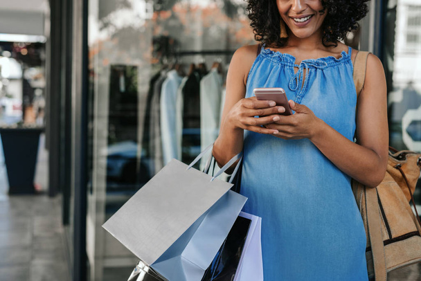 Closeup of a smiling young woman sending a text message on her cellphone while enjoying a day out shopping for clothing - Photo, image