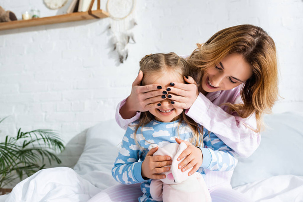 usmívající se a atraktivní matka zakrývající tvář dcery v pyžamu - Fotografie, Obrázek