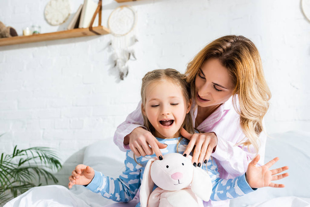 smiling mother showing soft toy to shocked daughter in bedroom  - Foto, afbeelding