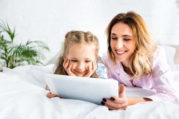 smiling mother and daughter using digital tablet in bedroom  - Fotografie, Obrázek