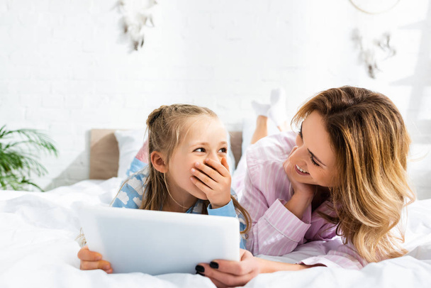 smiling mother looking at shocked daughter with digital tablet in bedroom  - Fotoğraf, Görsel
