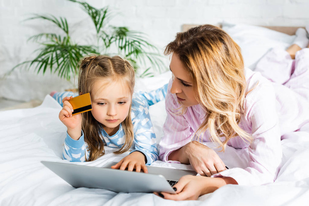 mother looking at laptop and daughter holding credit card in bedroom  - Foto, immagini
