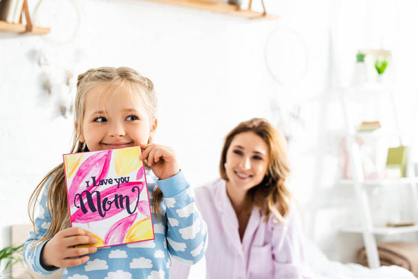 selective focus of daughter holding card with I love you mom lettering and smiling mother on background  - Photo, Image