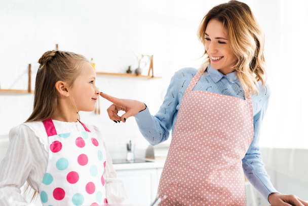 χαμογελαστή μητέρα αγγίζοντας τη μύτη της χαριτωμένης κόρης της στην κουζίνα  - Φωτογραφία, εικόνα