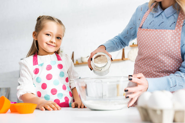 cropped view of mother adding flour to bowl and smiling daughter looking at camera  - Photo, Image