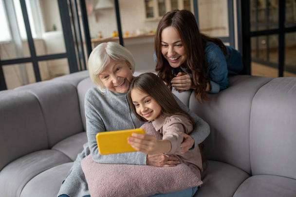 Happy girl, mom, grandmother taking pictures together at home on couch. - Foto, Imagem
