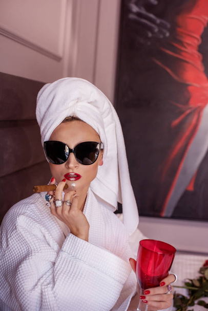 Stylish girl in a white bathrobe and towel wound on her head in black glasses with a cigar in her hands on the bed in the room. - Foto, Bild