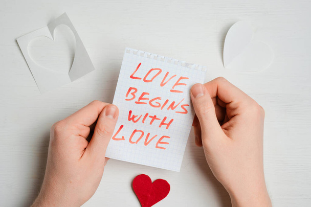 A hand holds a piece of paper with a red inscription - love begins with love - next to abstract hearts on a wooden white background. - Photo, Image