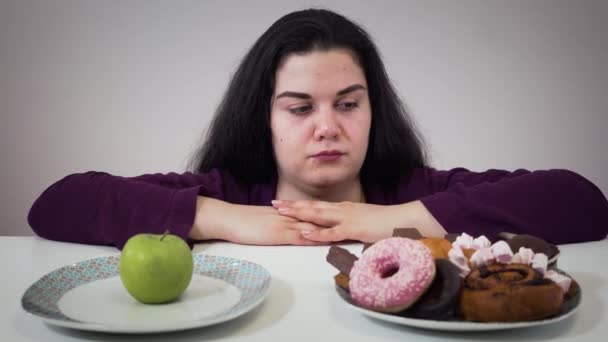 Fat Caucasian woman moving aside plate with apple and taking unhealthy sweets to eat. Portrait of smiling plump girl with overweight problem. Obesity, unhealthy lifestyle. - Footage, Video