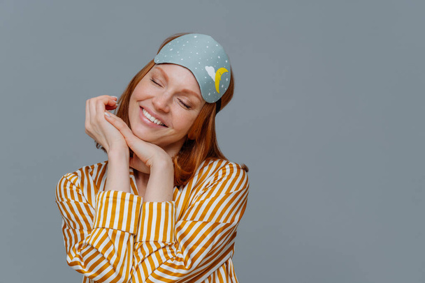 Portrait of good looking cheerful woman tilts head and smiles gently, imagines something pleasant, wears sleep mask or blindfold and pyjama, isolated over grey background, poses in cozy bedroom - 写真・画像
