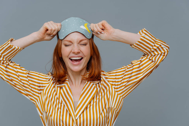 Overjoyed red haired Caucasian woman laughs positively, wears blindfold and yellow striped pajama, expresses good emotions, isolated on grey background. Good rest, bedtime and sleeping concept - Photo, Image