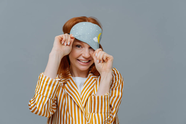 Playful woman with ginger hair looks from sleep mask, wears striped pajamas, has cheerful expression, poses over grey background, enjoys sleeping at home. Healthy sleep and relaxation concept - Photo, Image