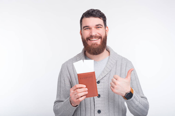 Fénykép fiatal jóképű férfi szakállal mutatja hüvelykujját fel, és kezében útlevél jegyekkel, készen áll az utazásra - Fotó, kép