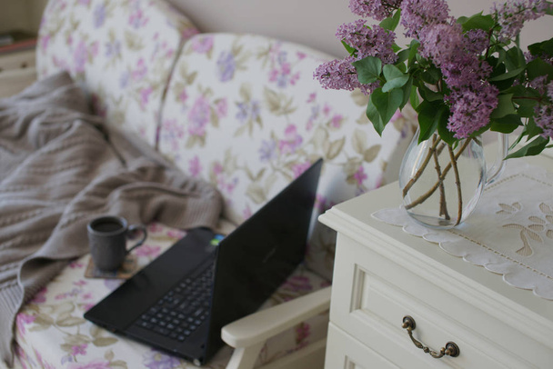 květinová pohovka s laptopem a šálkem čaje s dekou a šeříkovou kyticí na pozadí komodity - Fotografie, Obrázek