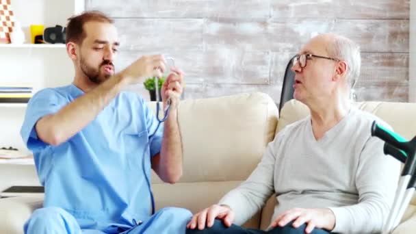 Revealing shot of young male nurse listening to old retired man heartbeat - Záběry, video