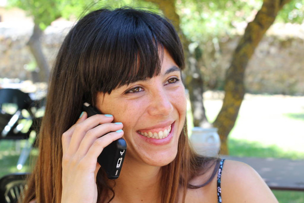 Портрет брюнетки красотки, сидящей в кафе и разговаривающей по телефону в летнем парке, размытый фон
   - Фото, изображение