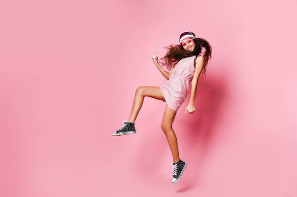 Teenager girl in a cap and pink striped dress is jumping joyfully shows a winner luck sign on a pink with copy space - Foto, Bild