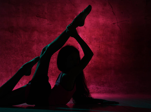 Silhouette of young slim girl doing gymnastic exercises lying on the floor stretching bent in lumbar holding legs up - Photo, Image