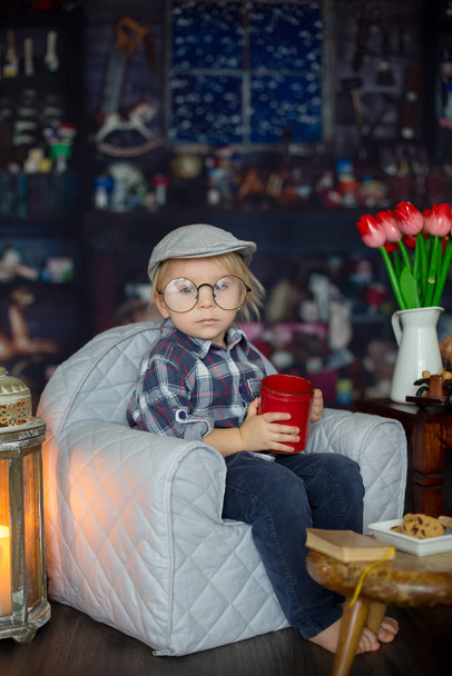 Sweet toddler boy, wearing glasses, reading a book and drinking  - Foto, Imagen