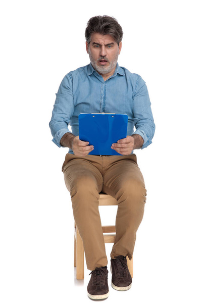 Σοκαρισμένος casual άνθρωπος ανάγνωση από ένα μπλε πρόχειρο και λαχανιάσει  - Φωτογραφία, εικόνα