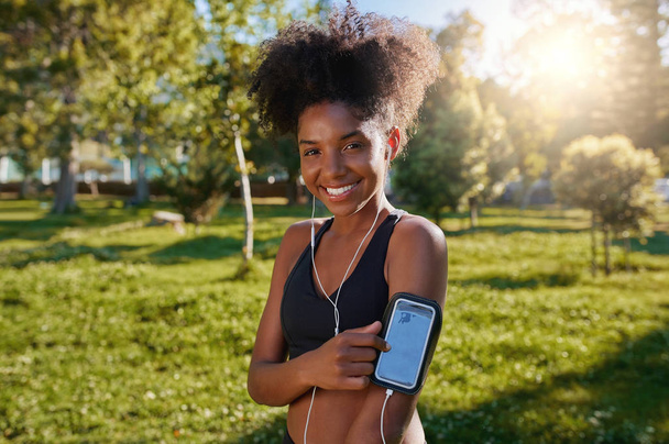 Portrait of a smiling african american young woman touching her mobile phone enjoying listening to music with earphones in the park - Φωτογραφία, εικόνα